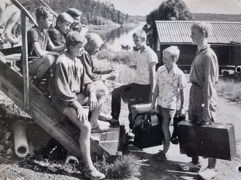 Pojkar med resväskor hälsas välkomna av lägerkompisar vid trappan nere vid vattnet